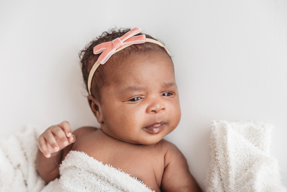 African American newborn Lily bare shoulder peeking out beneath a white blanket with pink bow at Reflection Images by Tracy Rowell in Florence SC