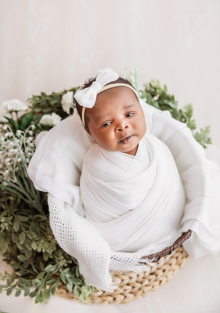 African American newborn Lily swaddled in white with a bow in a basket trimmed with greenery at Reflection Images by Tracy Rowell in Florence SC
