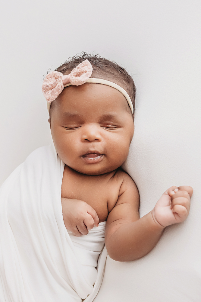 African American newborn Lily white wrap, pink bow, one arm peeking out in a relaxed pose at Reflection Images by Tracy Rowell in Florence SC