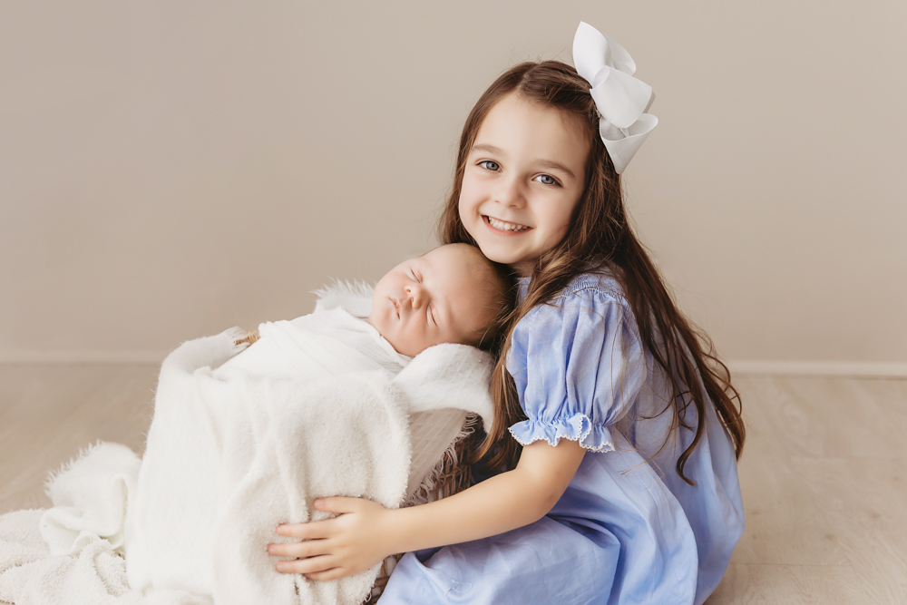 Big sister lying on white flokati rug beside Beau in a tan wrap, wearing blue dress with white bow at Reflection Images by Tracy Rowell in Florence SC