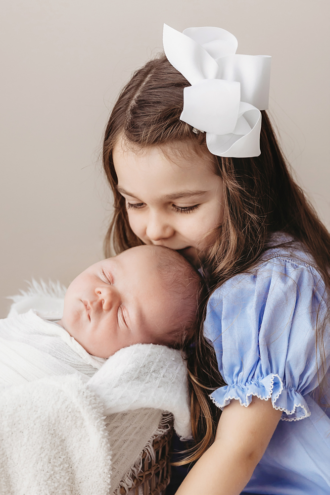 Big sister leans in to kiss newborn Beau swaddled in white wrap resting in a soft basket at Reflection Images by Tracy Rowell in Florence SC