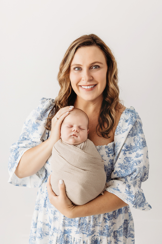 Mom smiling while holding swaddled Beau against white wall in blue toile dress at Reflection Images by Tracy Rowell in Florence SC