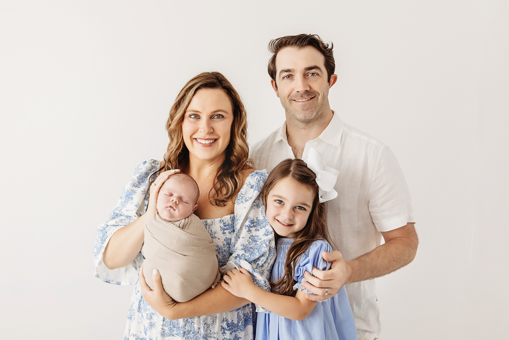 Family portrait with Beau swaddled in beige, held by mom in blue toile dress, dad and big sister in coordinated outfits at Reflection Images by Tracy Rowell in Florence SC