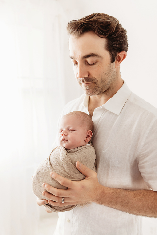 Dad in white shirt gazing at swaddled Beau under soft daylight near white curtains at Reflection Images by Tracy Rowell in Florence SC