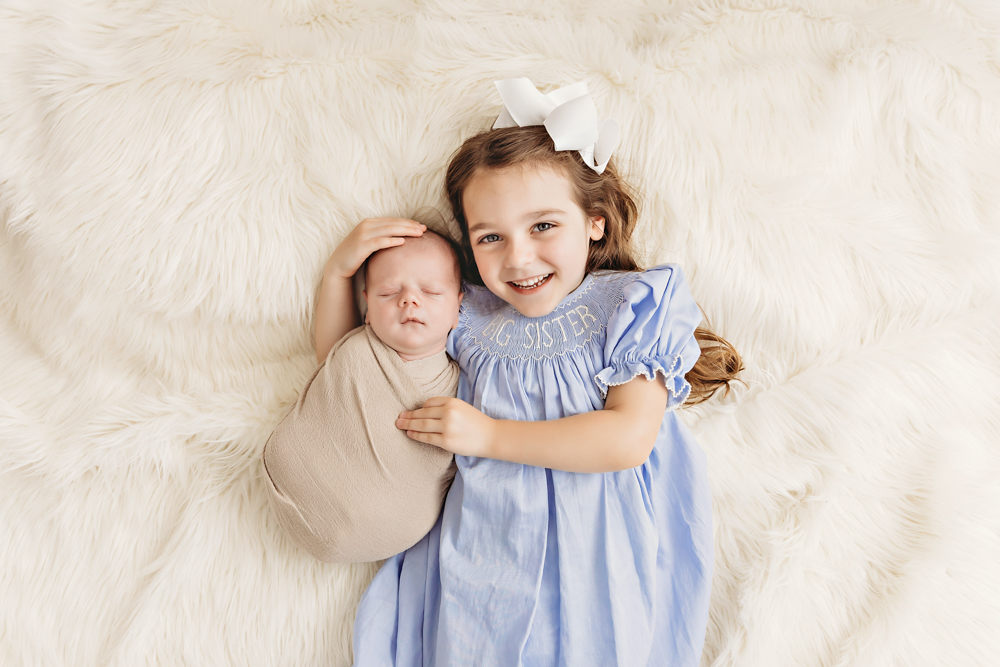 Beau swaddled in white blanket while big sister hugs and smiles proudly at Reflection Images by Tracy Rowell in Florence SC