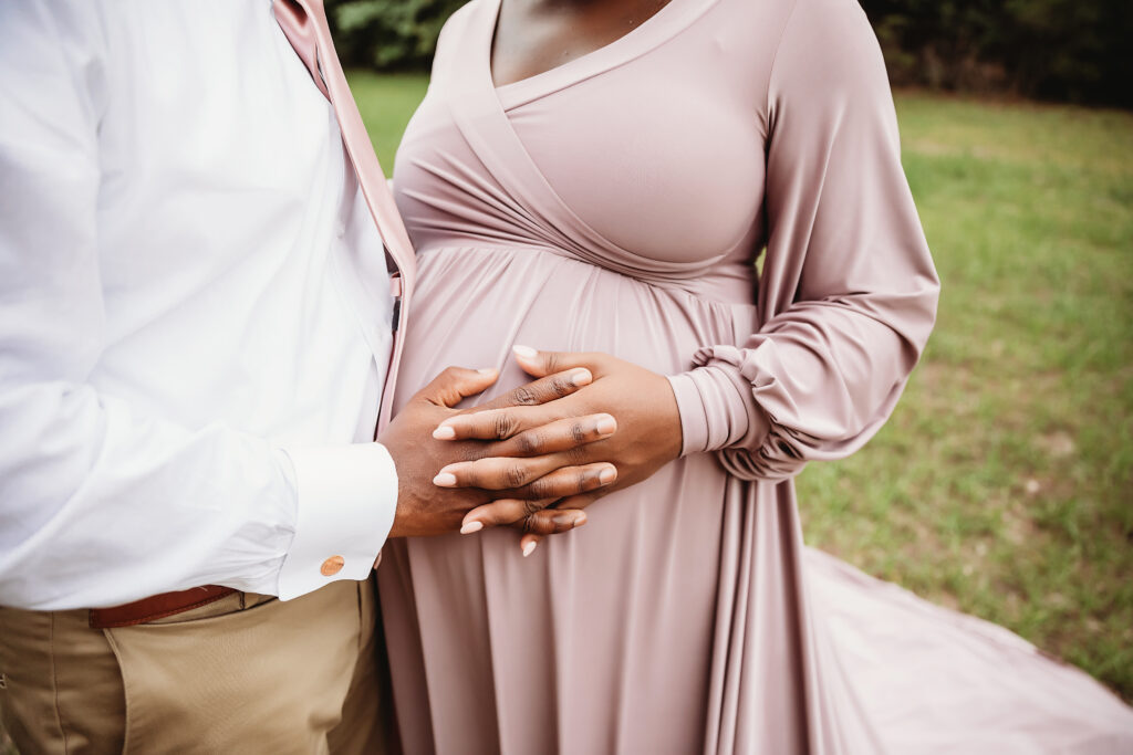 Close?up hands cradling baby bump, mauve sleeves, outdoor field in Florence SC