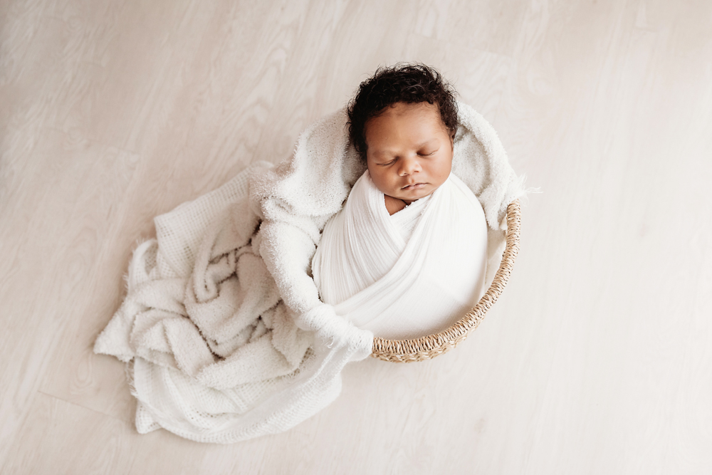 Braylen wrapped in white swaddle nestled in a wicker basket on a light wood floor at Reflection Images by Tracy Rowell in Florence SC