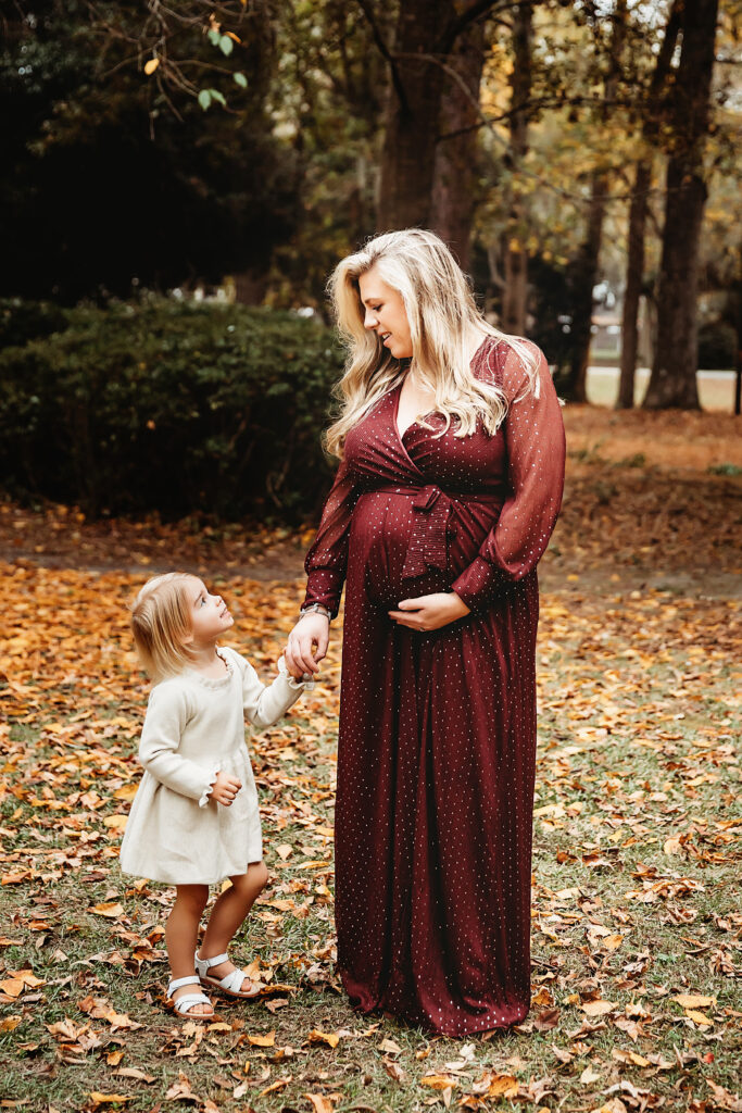 Pregnant mom in a burgundy dress holding hands with daughter in a cream dress among fall leaves at Reflection Images by Tracy Rowell in Florence SC