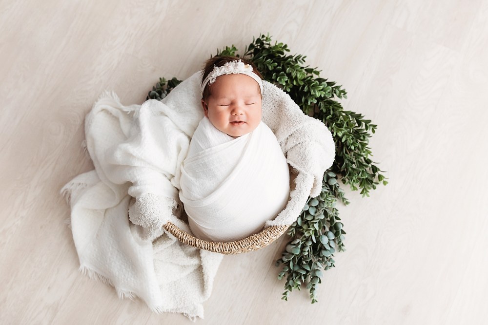 Overhead newborn swaddled in white, resting in basket with greenery at Reflection Images by Tracy Rowell in Florence SC.