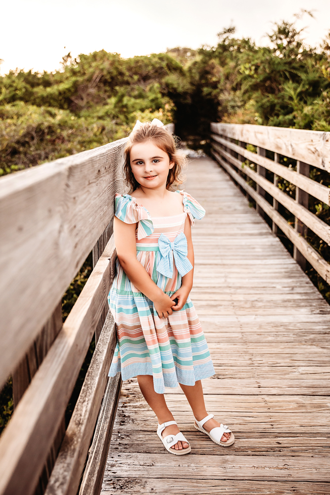 Stella in a pastel striped dress standing on a weathered beach boardwalk with greenery in the background at Reflection Images by Tracy Rowell in Florence SC