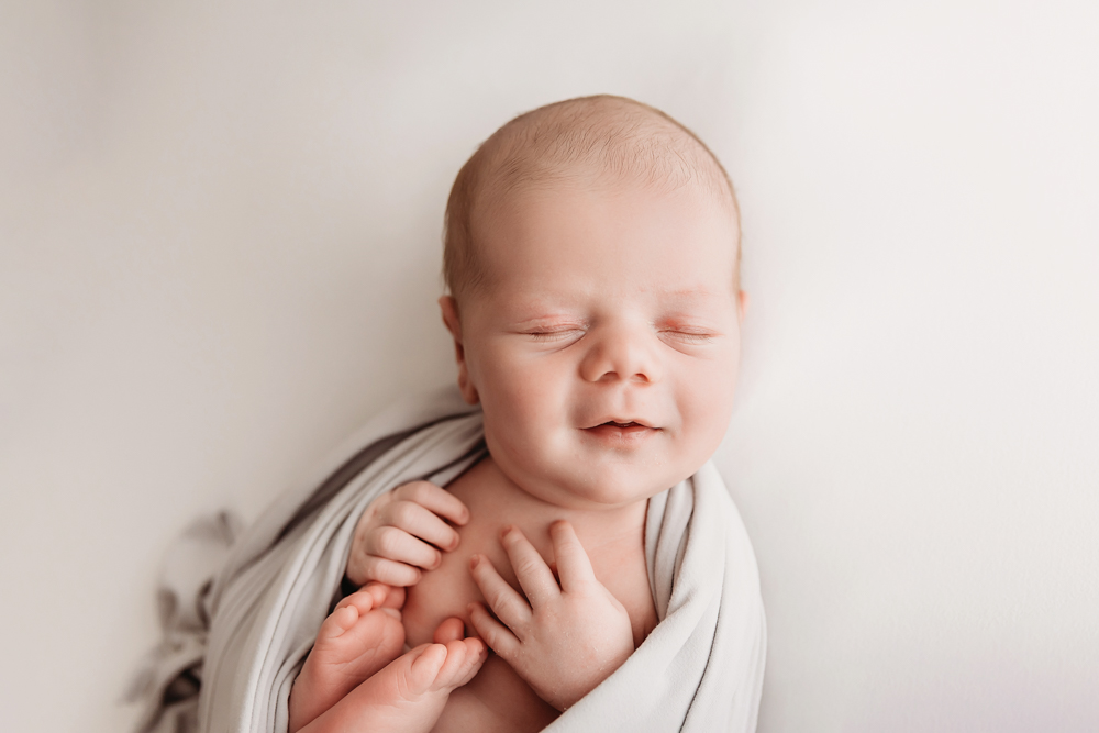 Beau swaddled in soft gray wrap with toes and fingers peeking out, smiling gently on white background at Reflection Images by Tracy Rowell in Florence SC