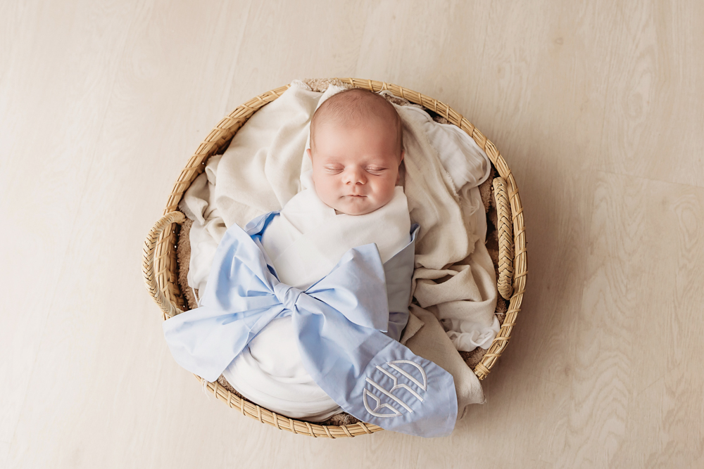 Overhead view of Beau swaddled in white wrap with oversized monogrammed blue bow, nestled in basket on wood floor at Reflection Images by Tracy Rowell in Florence SC
