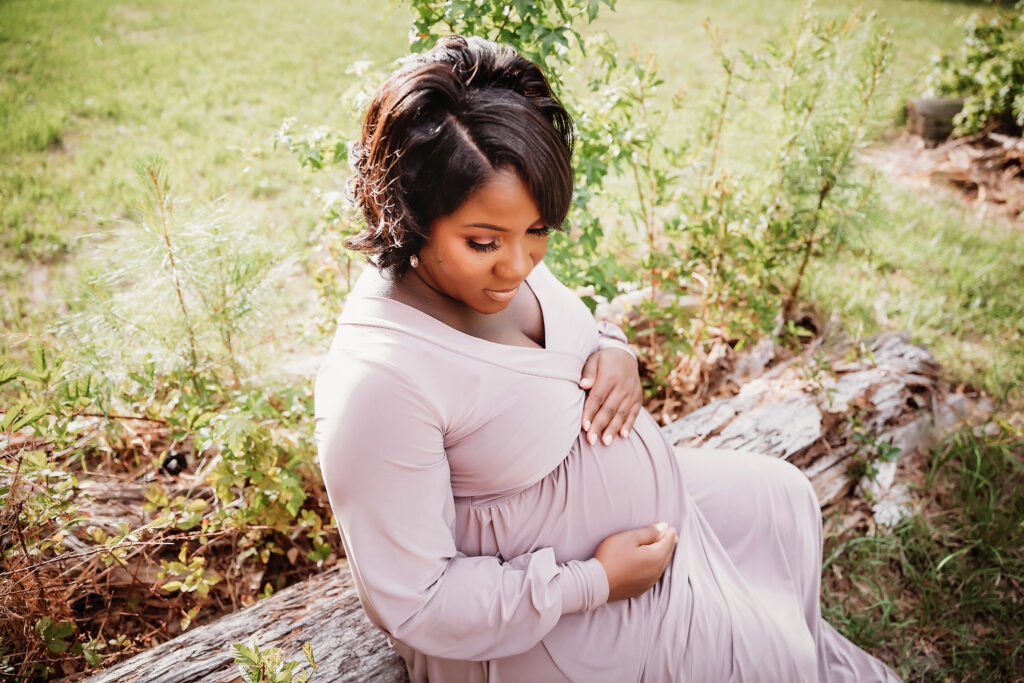 Chelsea seated on a log gently holding her bump, greenery surrounding, Florence SC.