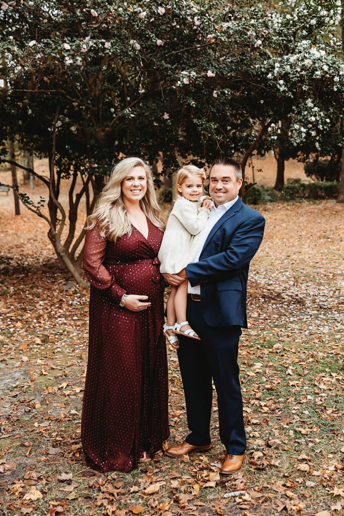 Expecting couple with daughter posed for a family portrait under flowering trees in a park in Florence SC, mom in burgundy, dad in navy suit