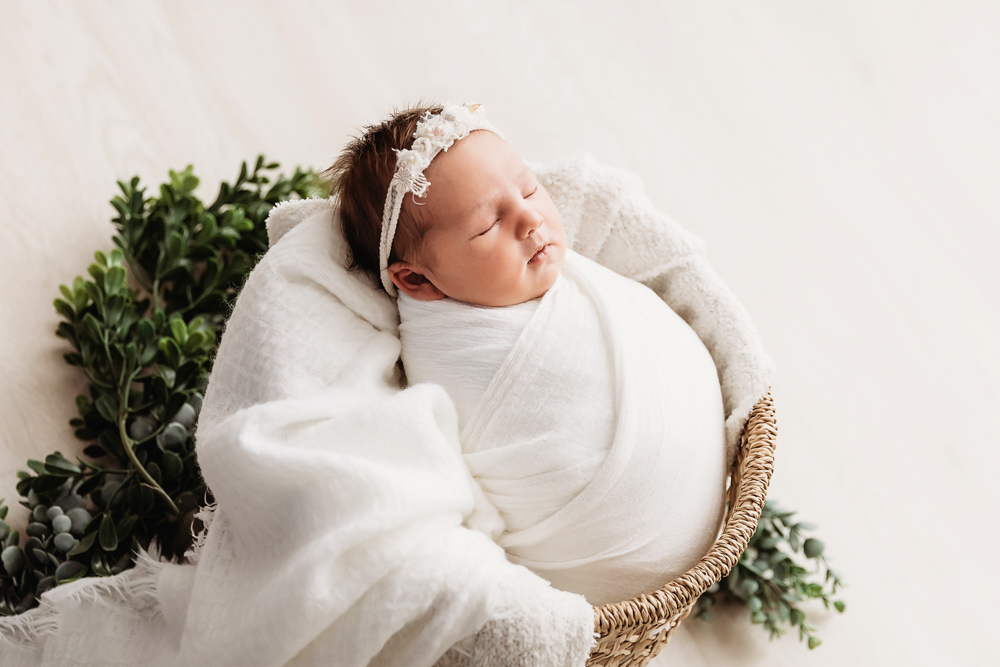 Side-angle view of newborn swaddled in white, in basket styled with greenery at Reflection Images by Tracy Rowell in Florence SC.