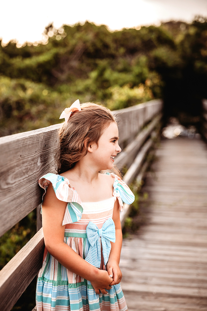 Stella smiling sideways with a white bow and ruffle dress on a wooden boardwalk surrounded by greenery at Reflection Images by Tracy Rowell in Florence SC