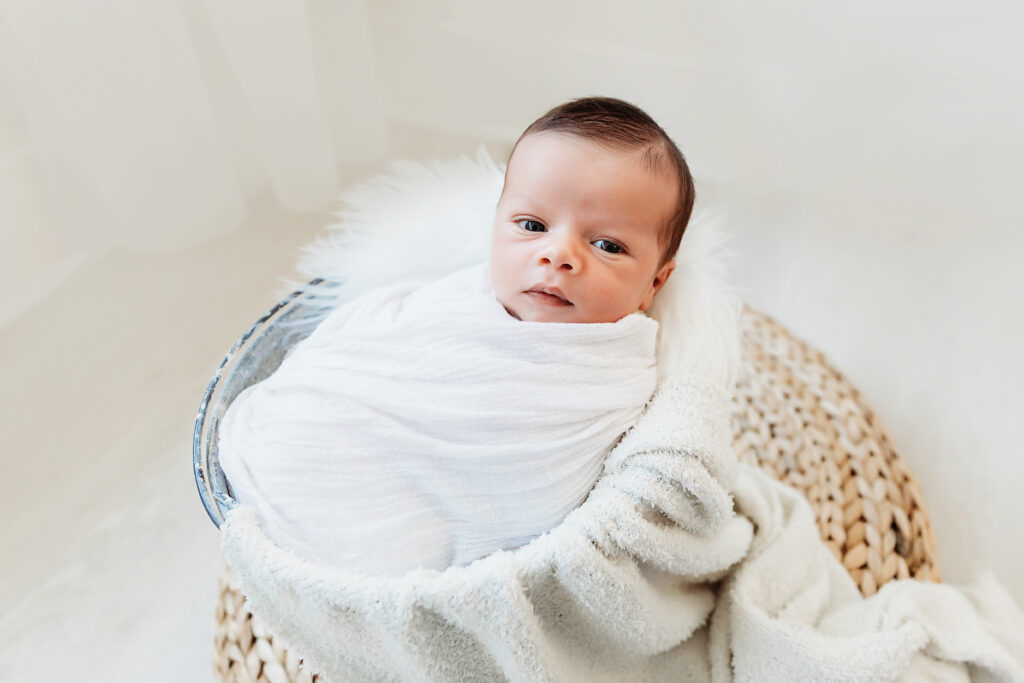 Newborn baby in a white wrap wide-eyed in a wicker basket with fluffy white pillow at Reflection Images by Tracy Rowell in Florence SC