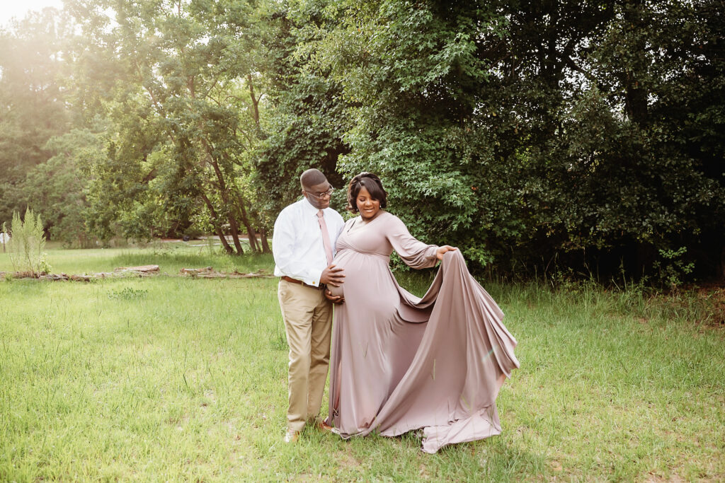Couple smiling together, mother-to-be holding lavender gown while father holds her bump in green field at Reflection Images by Tracy Rowell in Florence SC.