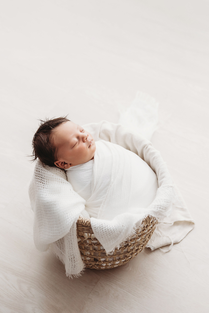 Newborn Liston swaddled in white, sleeping in a woven basket on a light wood floor at Reflection Images by Tracy Rowell in Florence SC