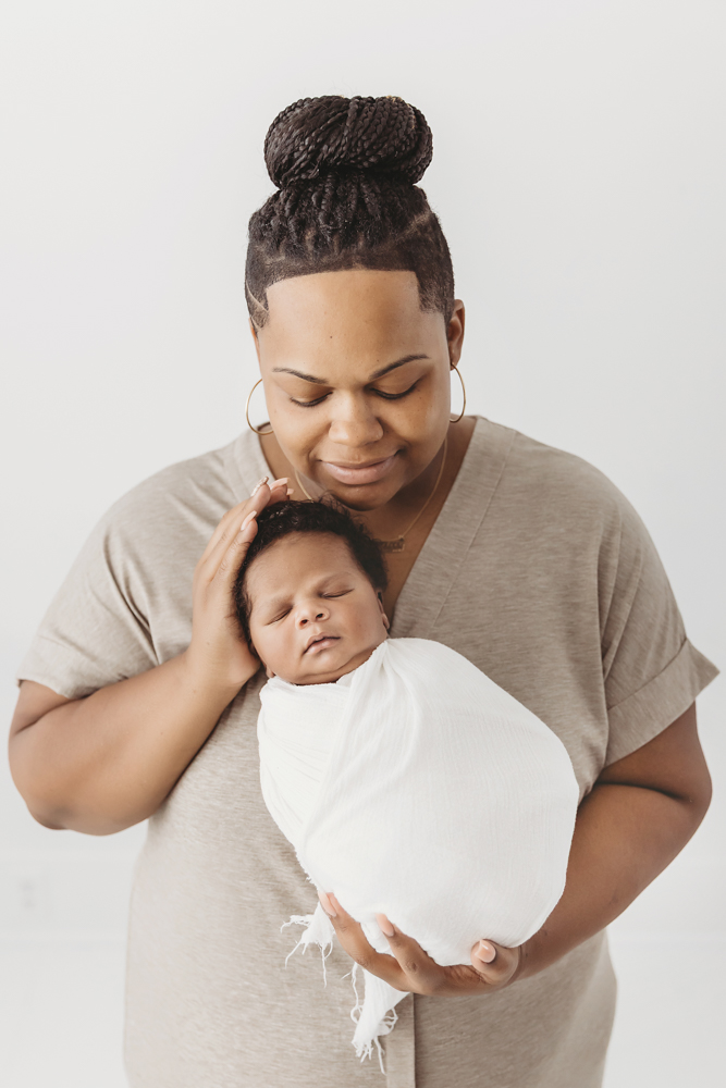 Braylen swaddled in white with moms hand resting on his head, set against a bright studio backdrop at Reflection Images by Tracy Rowell in Florence SC