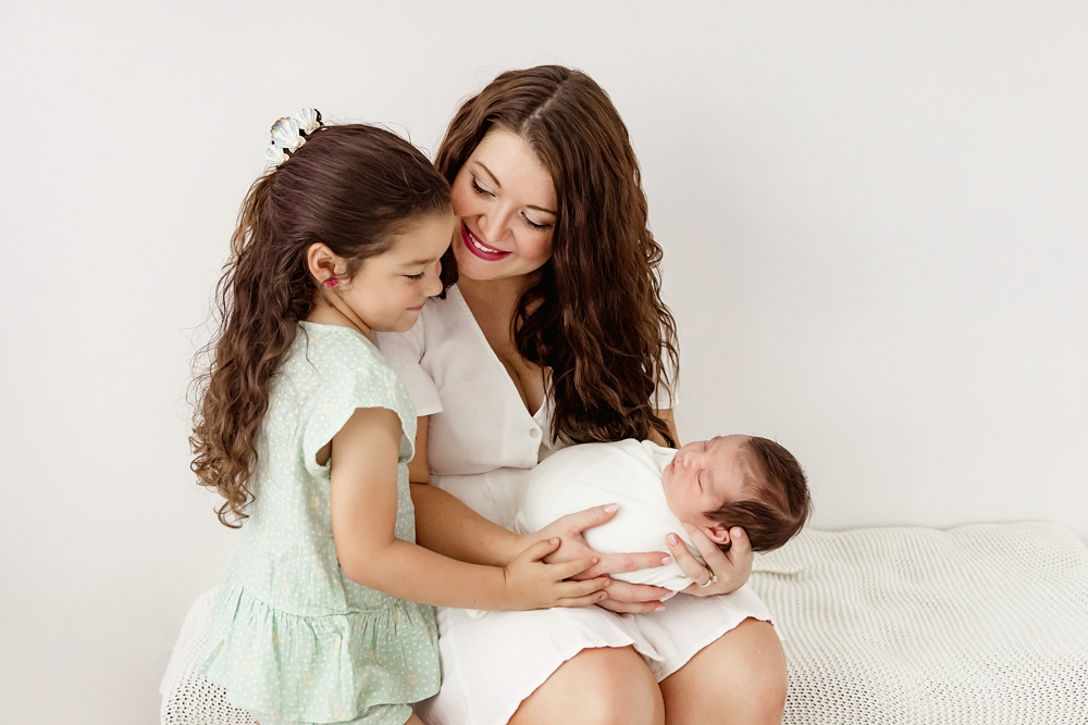 Older sister smiling while mom holds swaddled newborn Finn, with gentle sibling interaction at Reflection Images by Tracy Rowell in Florence SC