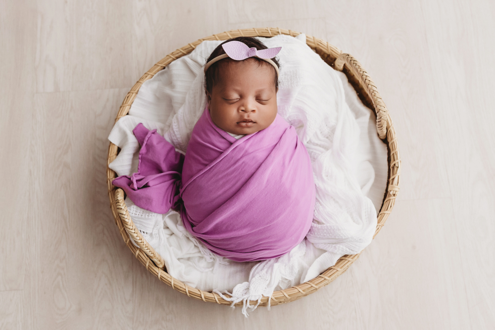 Baby Harlem swaddled in a purple wrap with matching bow, asleep in a round basket on white layers at Reflection Images by Tracy Rowell in Florence SC