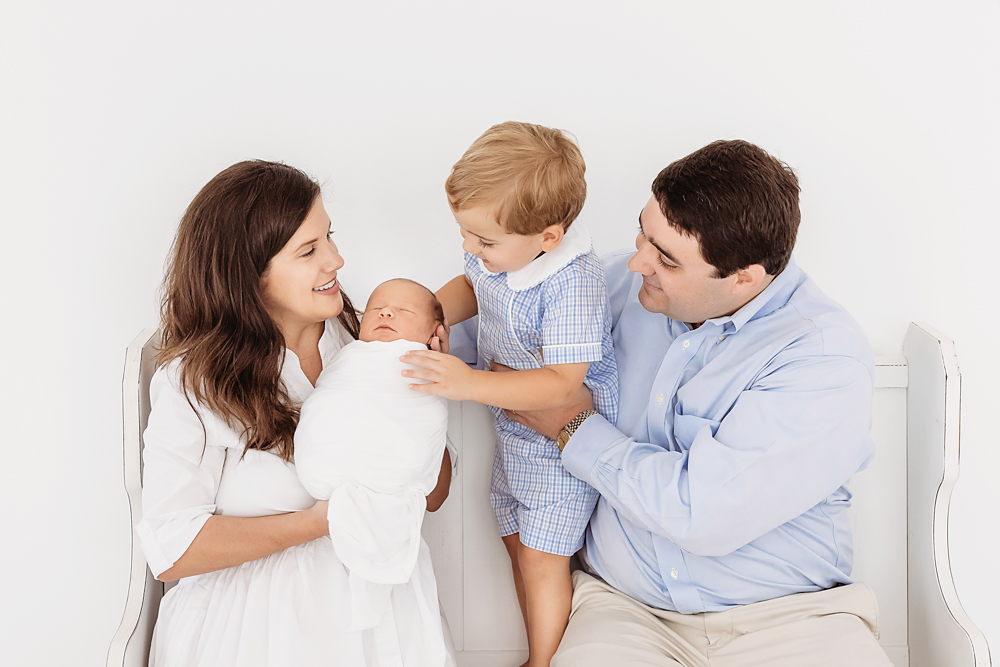 Tommy leaning in to kiss newborn brother Harry swaddled in soft white wrap with family at Reflection Images by Tracy Rowell in Florence SC