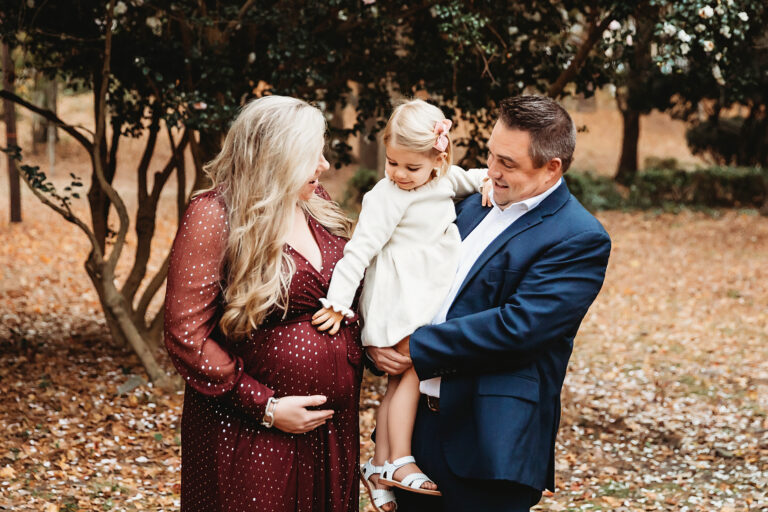 Expecting mom in burgundy sharing a laugh with dad in navy while daughter with pink bow looks on at Reflection Images by Tracy Rowell in Florence SC