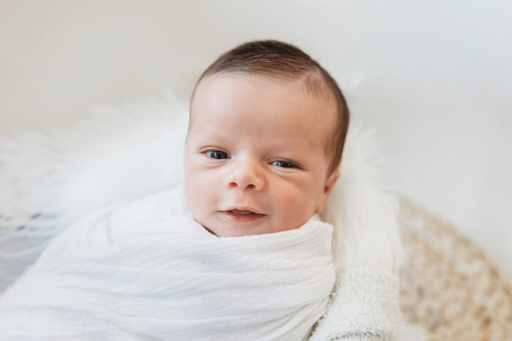 Newborn baby smiling while wrapped in a white blanket in a wicker basket on flokati at Reflection Images by Tracy Rowell in Florence SC