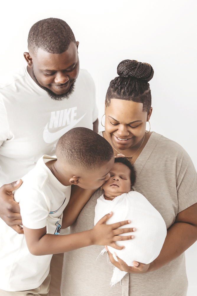 Family moment with big brother kissing baby Braylen in a white wrap, surrounded by smiles at Reflection Images by Tracy Rowell in Florence SC