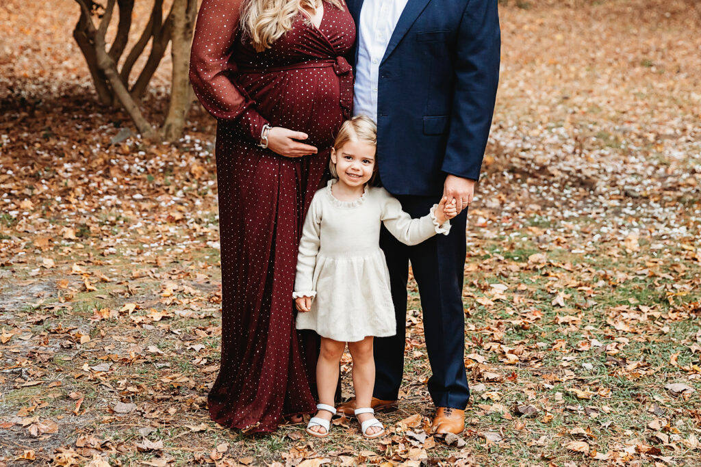 Little girl smiling while holding her parents' hands on a blanket of fall leaves during maternity session at Reflection Images by Tracy Rowell in Florence SC