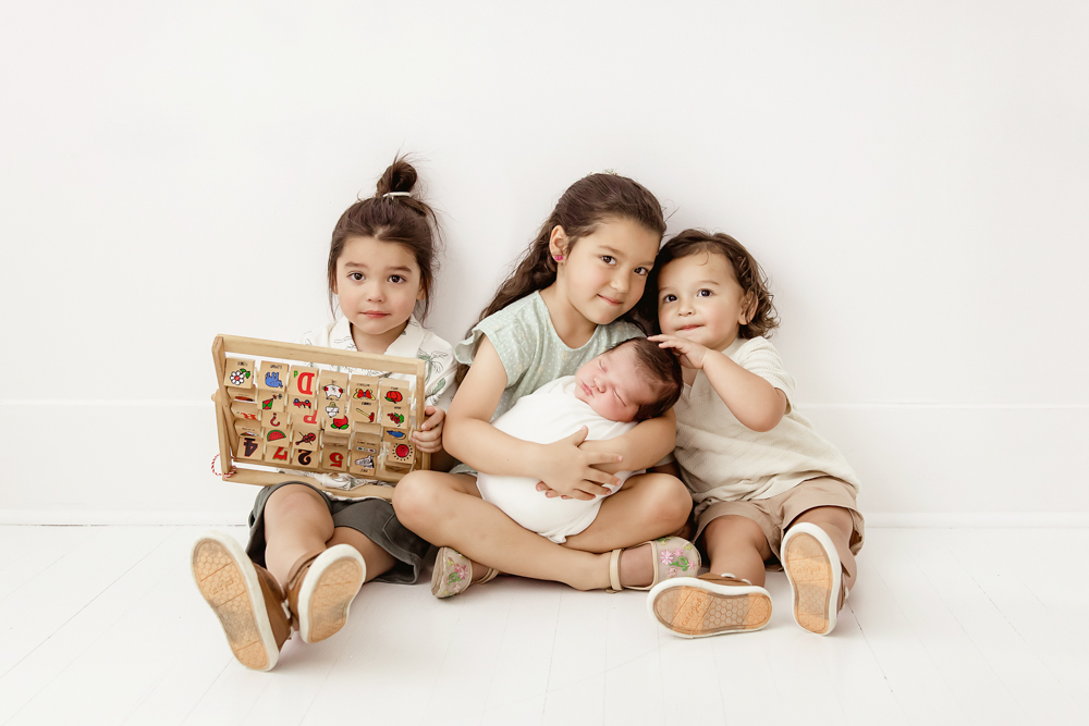All siblings seated on white floor with baby Finn in arms, natural expressions and soft light at Reflection Images by Tracy Rowell in Florence SC