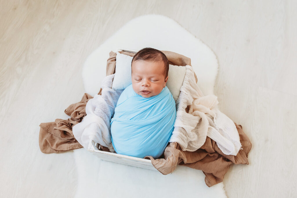 Top-down view of a newborn in a bright blue wrap, resting in a wooden crate on white flokati at Reflection Images by Tracy Rowell in Florence SC