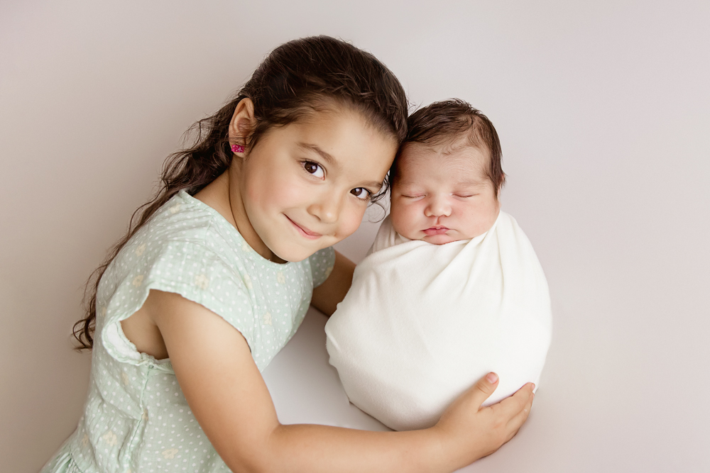 Big sister hugging baby Finn swaddled in white against a simple studio backdrop at Reflection Images by Tracy Rowell in Florence SC