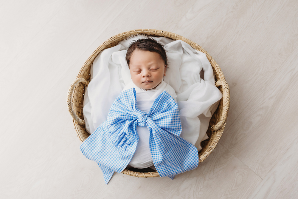 Liston swaddled in white with a personalized blue gingham bow in a basket on light wood floor at Reflection Images by Tracy Rowell in Florence SC