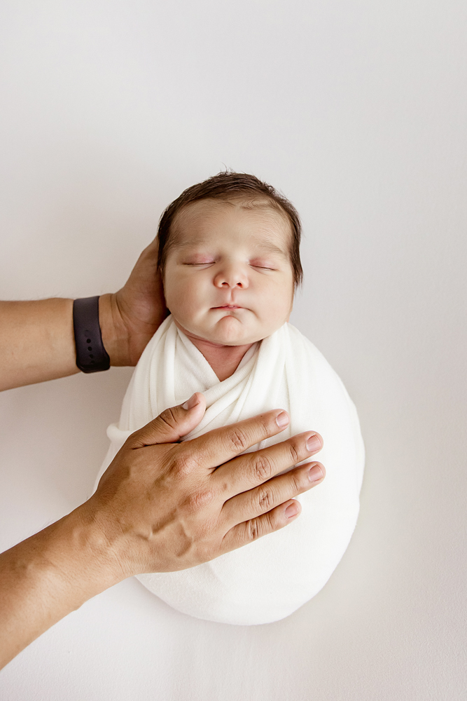 Close-up of hands gently holding baby Finn in white wrap during newborn session at Reflection Images by Tracy Rowell in Florence SC