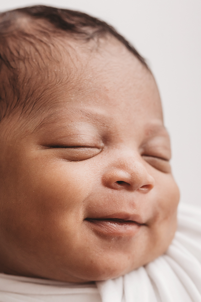 Harlem smiling in her sleep, close-up with dimples showing while swaddled in white wrap at Reflection Images by Tracy Rowell in Florence SC