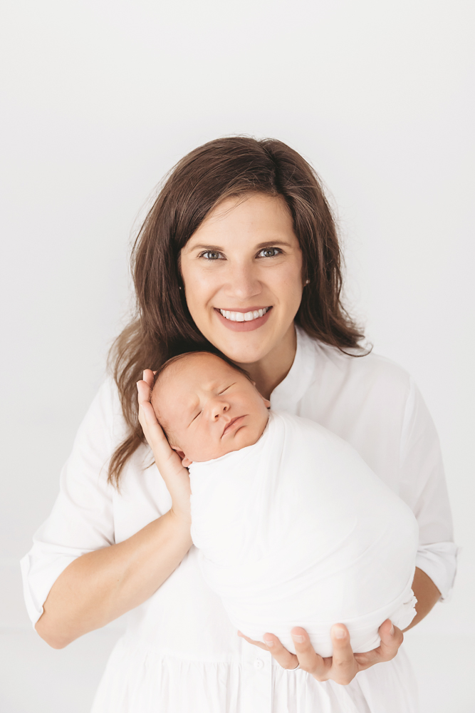Eliza smiling while holding swaddled Harry close in front of a white backdrop at Reflection Images by Tracy Rowell in Florence SC