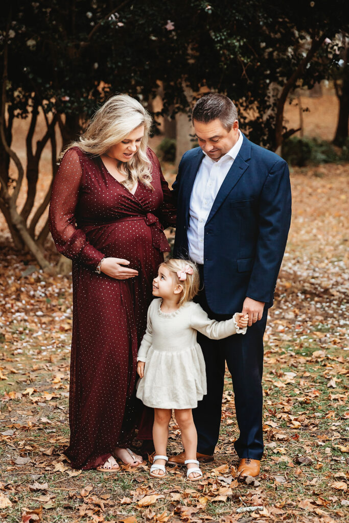 Maternity photo of parents looking lovingly at daughter in cream dress, fall leaves and flowering trees in the background in Florence SC
