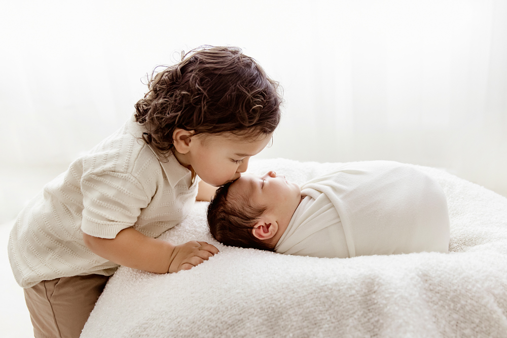 Toddler brother kissing swaddled newborn on forehead, natural light setting at Reflection Images by Tracy Rowell in Florence SC