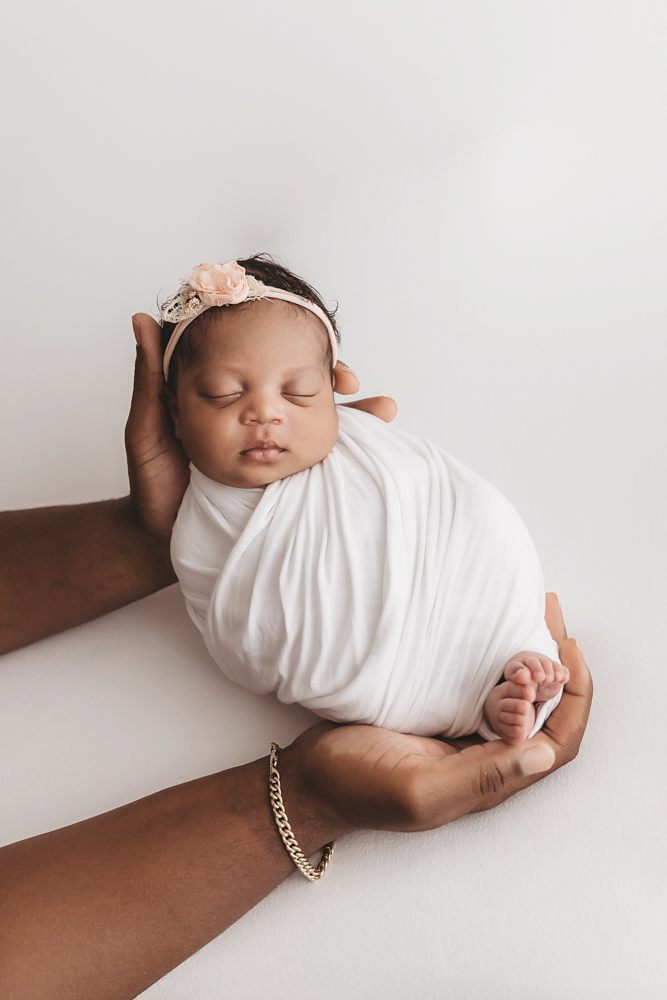 Harlem swaddled in white wrap with peach floral headband, resting in dads hands against a soft white backdrop at Reflection Images by Tracy Rowell in Florence SC