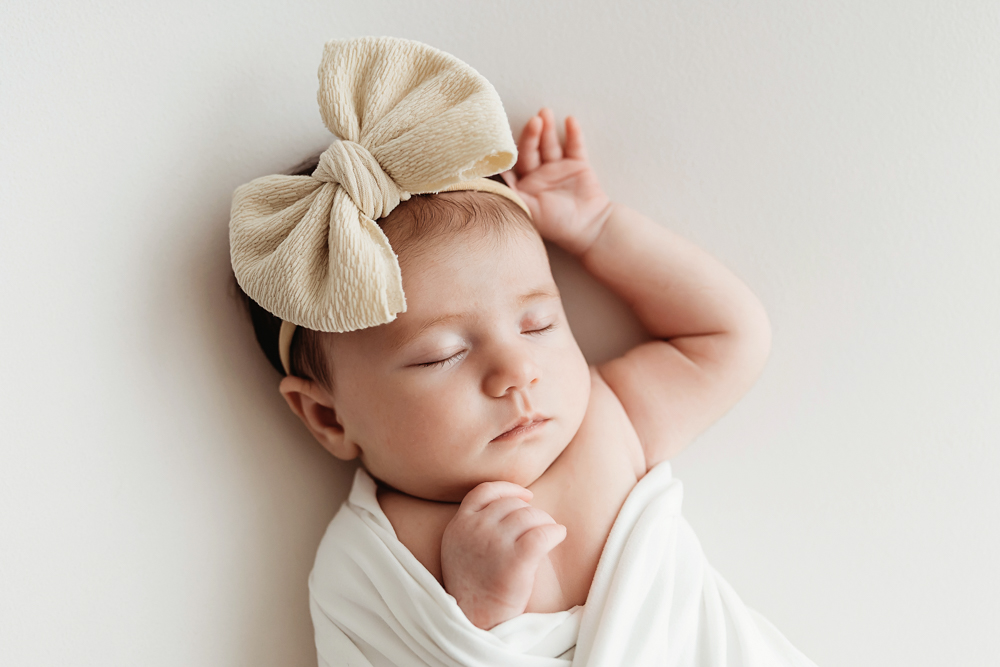 Close-up of Mary Katherine sleeping in a white wrap with an oversized beige bow headband on a soft studio backdrop at Reflection Images by Tracy Rowell in Florence SC