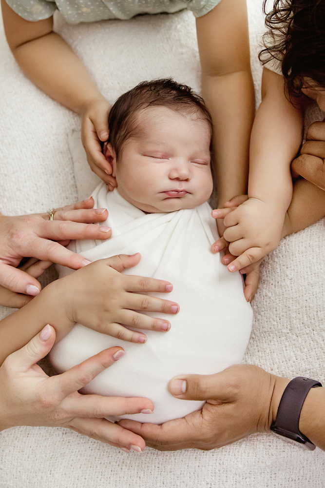 Group of sibling and parent hands resting on baby Finn wrapped in white at Reflection Images by Tracy Rowell in Florence SC