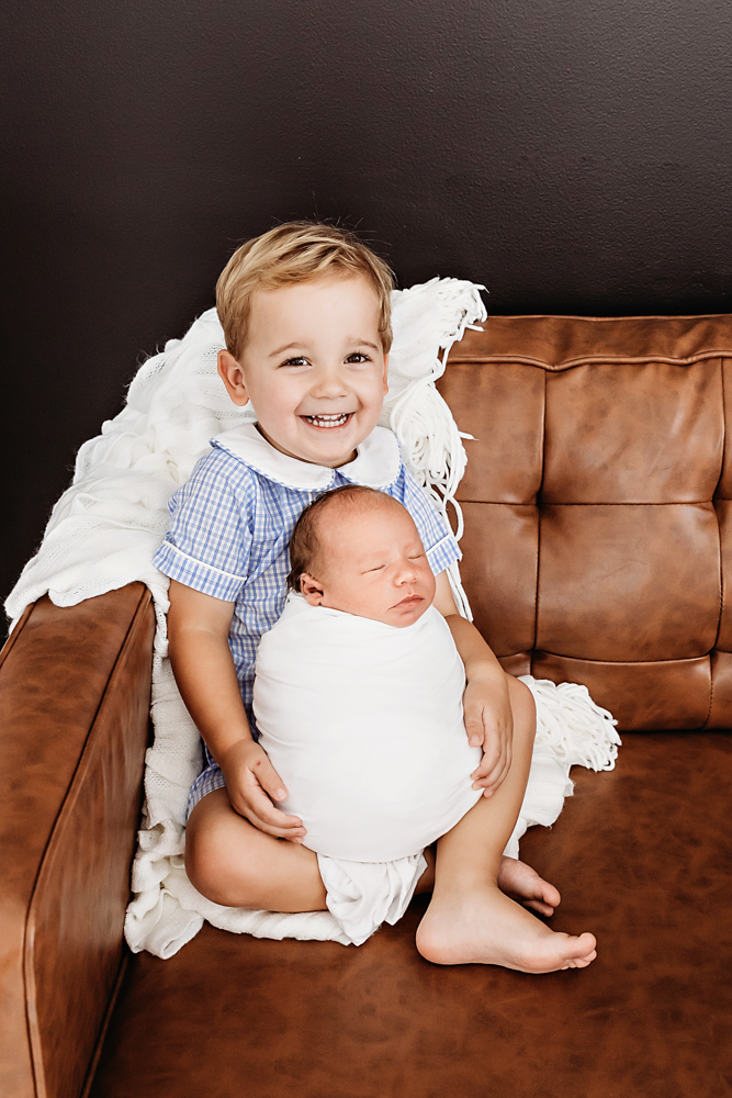 Tommy sitting on brown leather couch holding swaddled baby brother Harry with a big smile at Reflection Images by Tracy Rowell in Florence SC