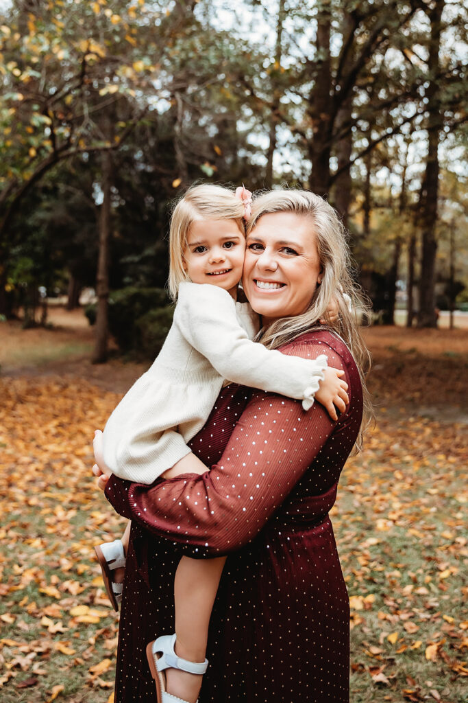 Expecting mom in burgundy dress holding daughter with pink bow and smiling under golden fall trees in Florence SC