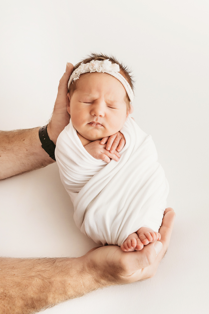 Baby swaddled in white wrap, cradled securely in fathers hands at Reflection Images by Tracy Rowell in Florence SC.
