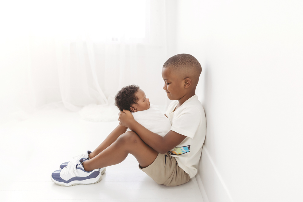 Big brother sitting against a white wall, carefully holding swaddled newborn Braylen in a heartwarming sibling moment at Reflection Images by Tracy Rowell in Florence SC