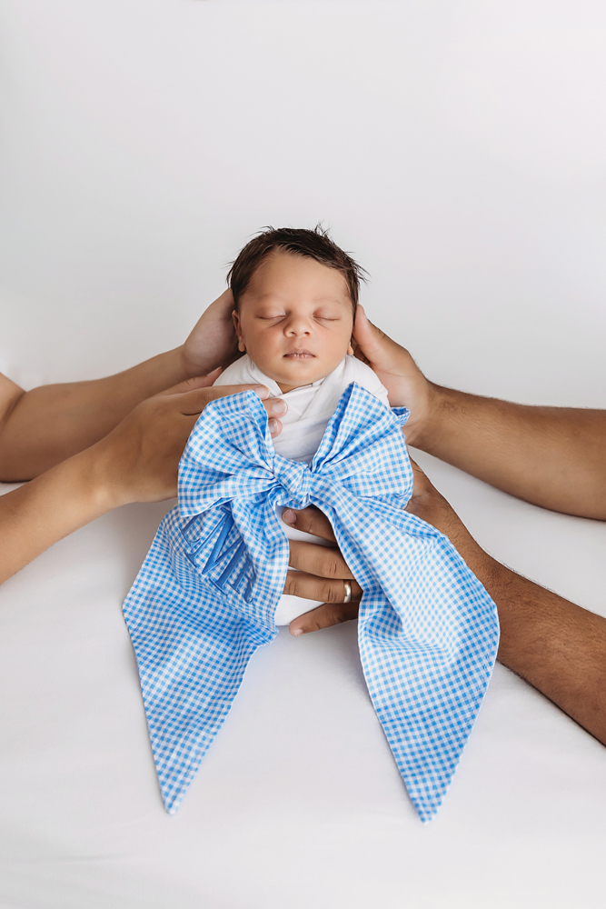 Family hands gently cradle newborn Liston wrapped in white with a large blue gingham bow at Reflection Images by Tracy Rowell in Florence SC