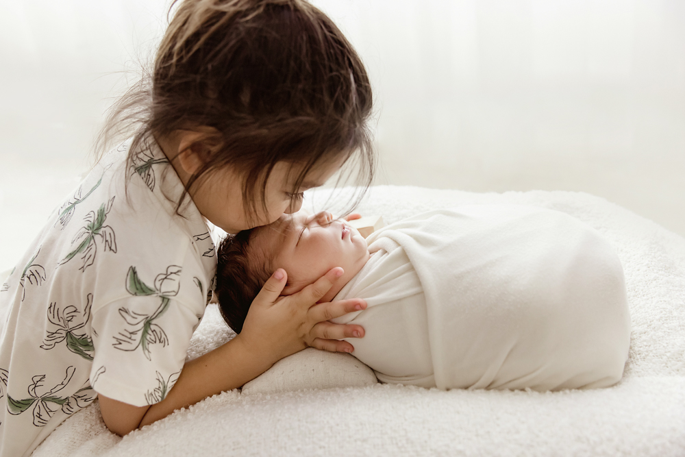 Older sibling leaning in to kiss newborn Finn wrapped in white, overhead angle at Reflection Images by Tracy Rowell in Florence SC