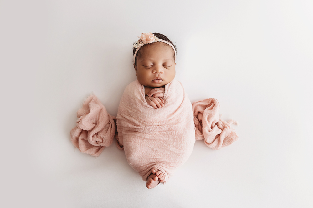 Top-down view of Harlem swaddled in a blush pink wrap with matching floral headband, asleep on a white studio backdrop at Reflection Images by Tracy Rowell in Florence SC
